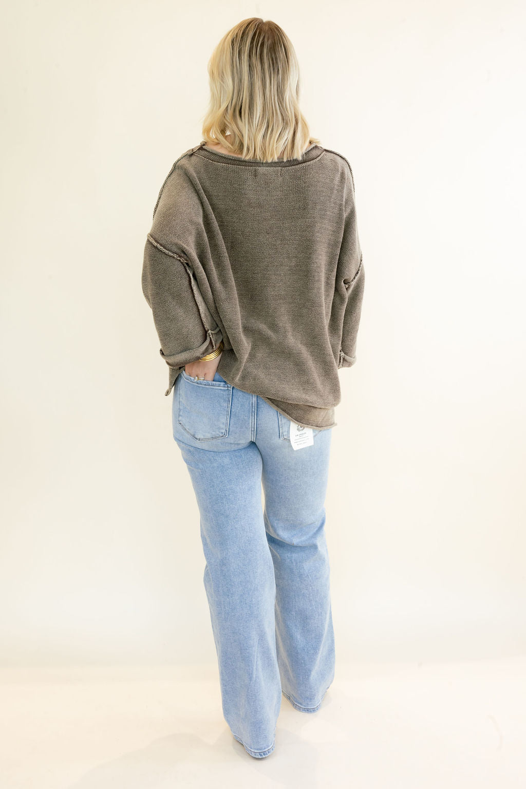 A woman shown from the back wearing a loose, oversized dark brown knit top and light-wash blue jeans against a plain white studio background.