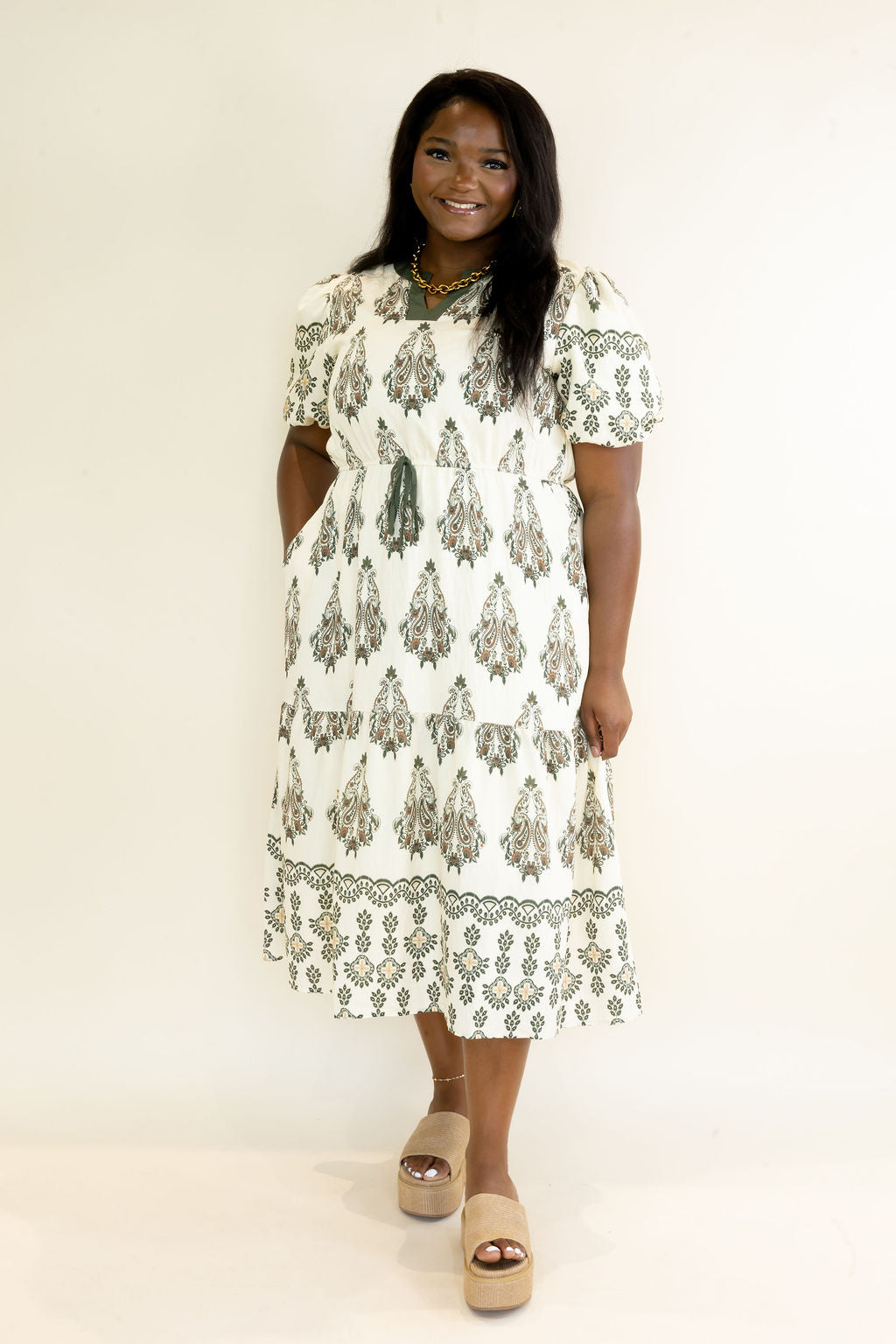 Full front view of woman modeling ivory and olive green botanical print midi dress with puff sleeves, side pockets, tiered skirt, and midi length