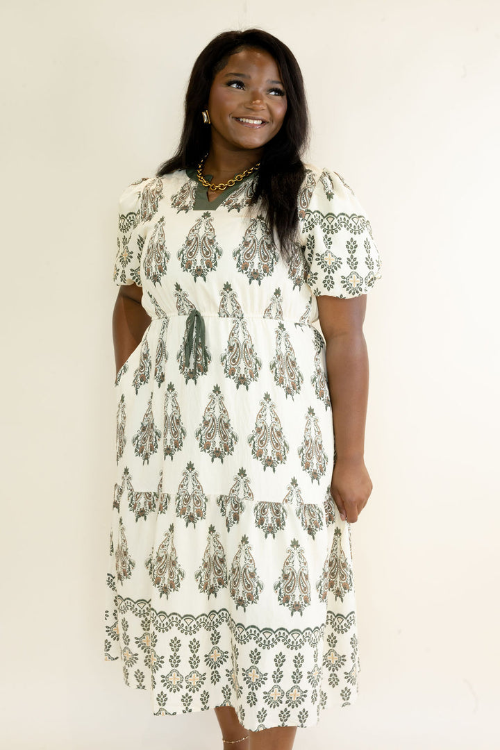 Woman modeling ivory midi dress with olive green botanical print, puff short sleeves, tiered skirt, and decorative border print