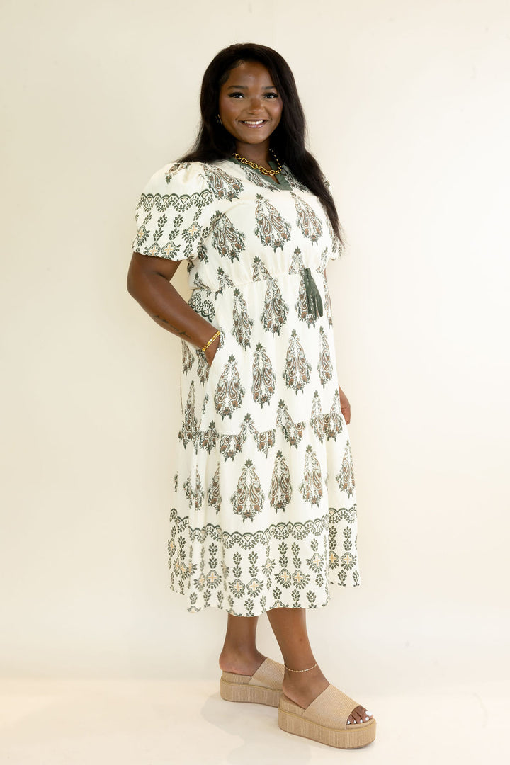 Three-quarter view of woman wearing ivory midi dress with olive green botanical print, puff sleeves, tiered silhouette, and neutral platform shoes