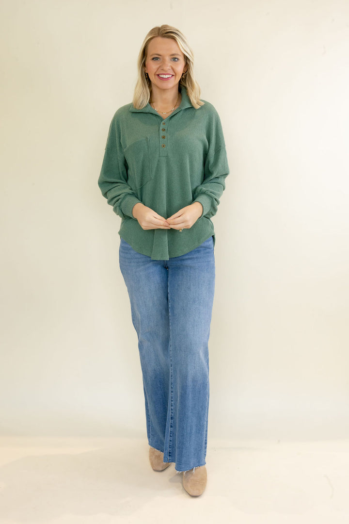 A woman modeling a sage green long-sleeve polo-style top with blue jeans and beige shoes, standing against a plain off-white studio background.