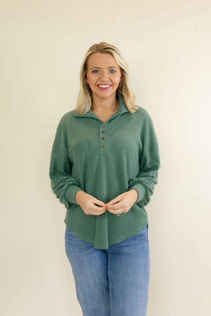 A smiling blonde woman wearing a sage green ribbed knit polo-style sweater with a button placket and blue jeans, standing against a plain off-white wall.