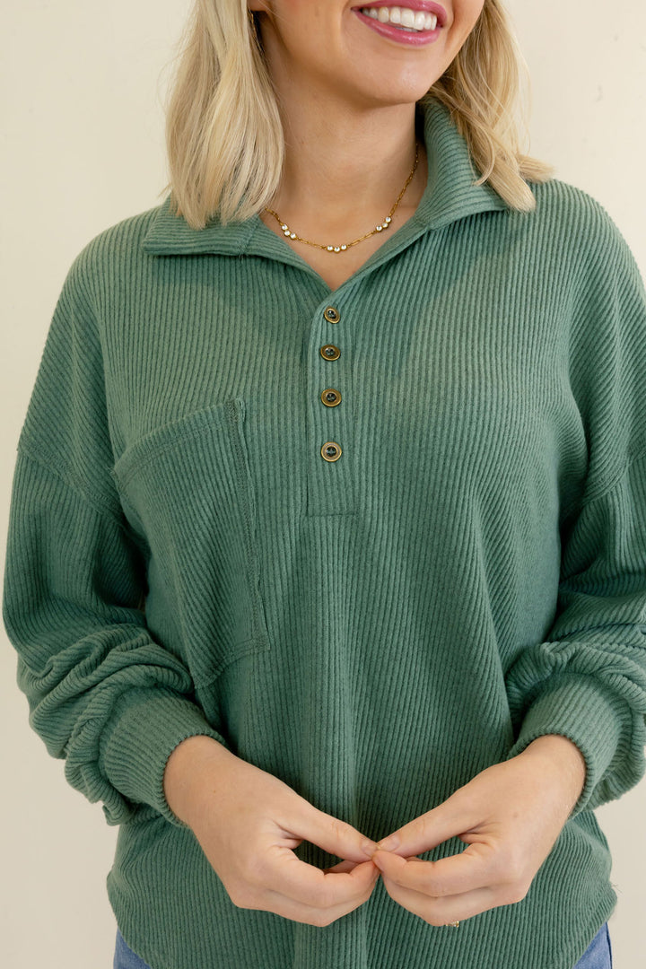 A woman wearing a sage green ribbed knit polo-style sweater with a partial button placket and a chest pocket, shown from the shoulders to the waist against a plain light beige background.