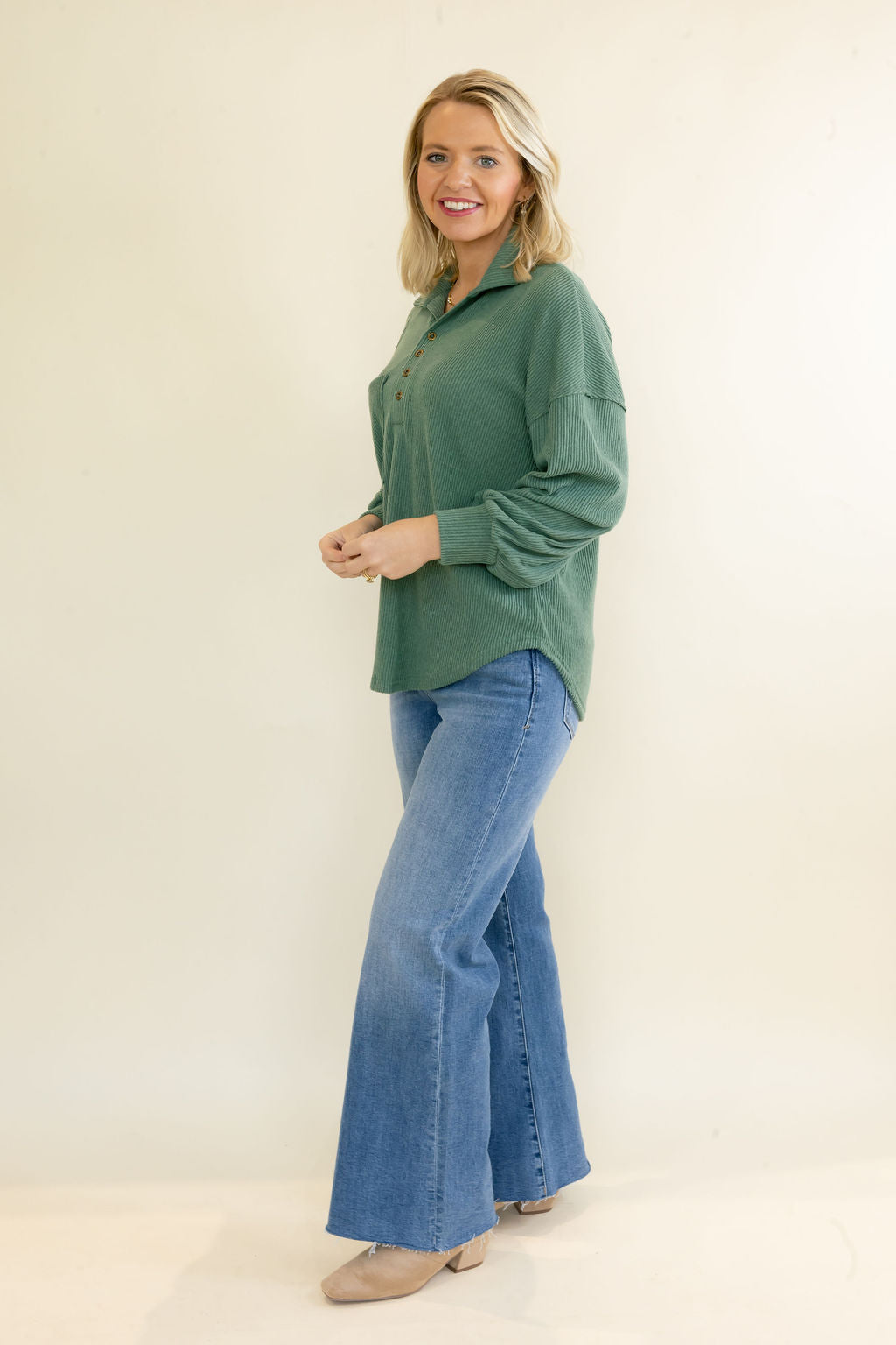 A woman modeling a sage green long-sleeve button-up shirt and blue flared jeans against a plain cream studio background.