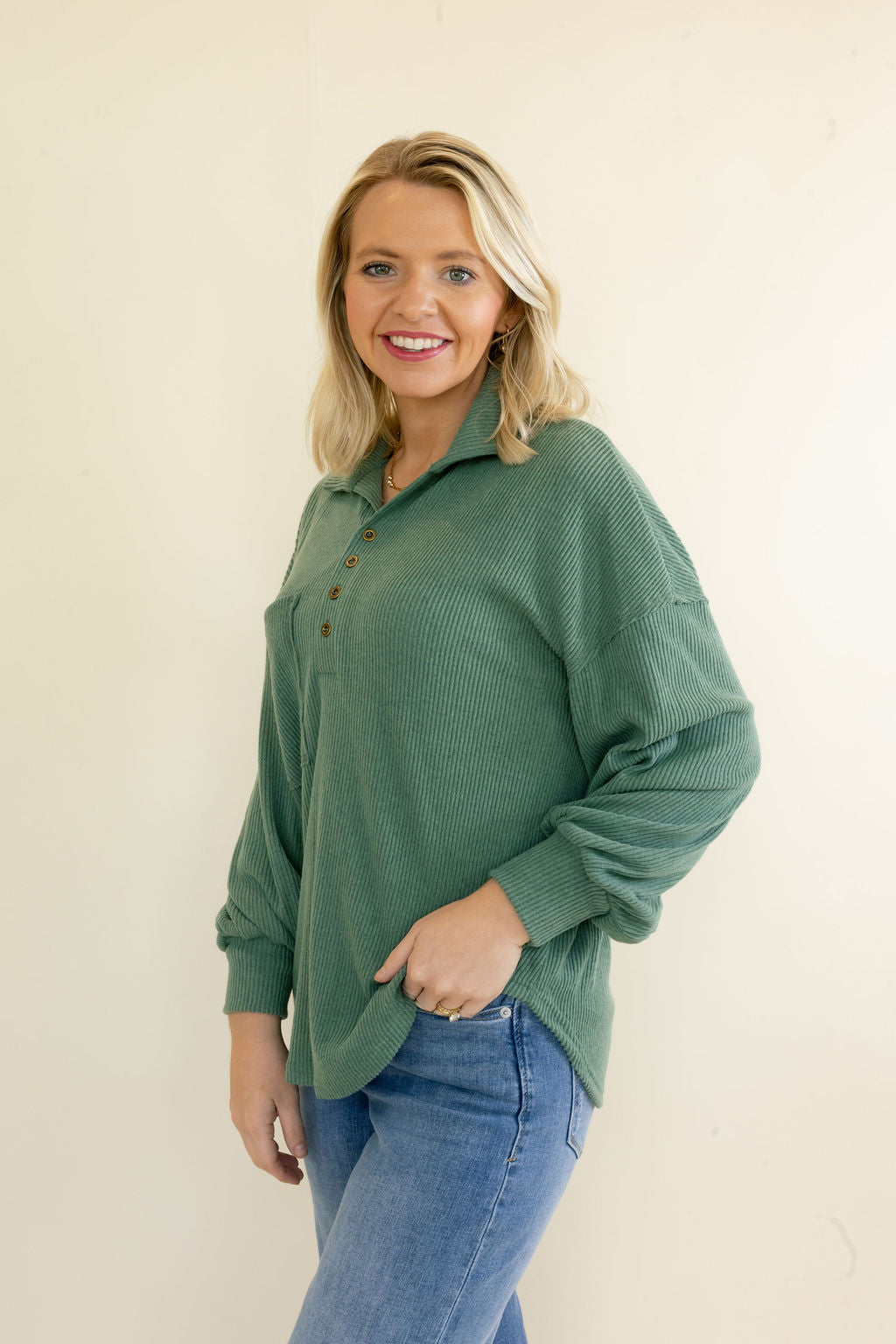 Woman modeling sage green ribbed collared pullover top with button neckline, drop shoulders, long sleeves, and relaxed fit styled with blue jeans