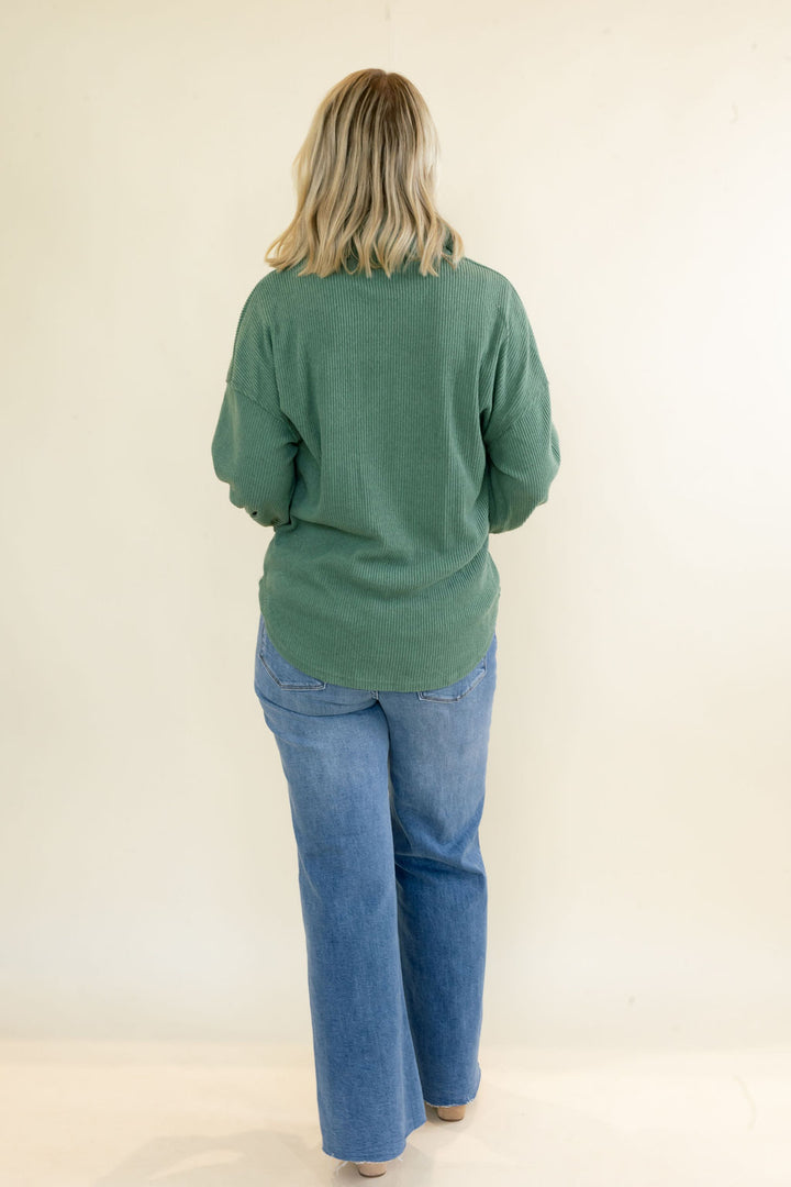A woman viewed from behind wearing a sage green long-sleeve top and blue jeans, standing against a plain off-white background.