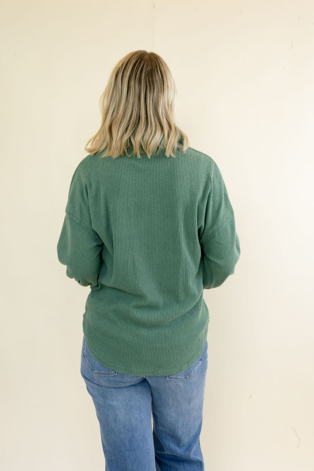 A woman with blonde hair is shown from behind wearing a sage green ribbed knit sweater and blue jeans against a plain off-white wall.