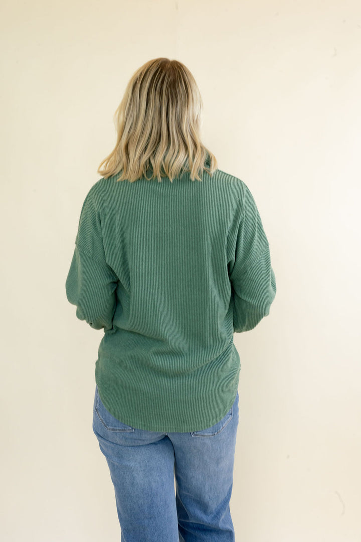 A woman with blonde hair is shown from behind wearing a sage green ribbed knit sweater and blue jeans against a plain off-white wall.