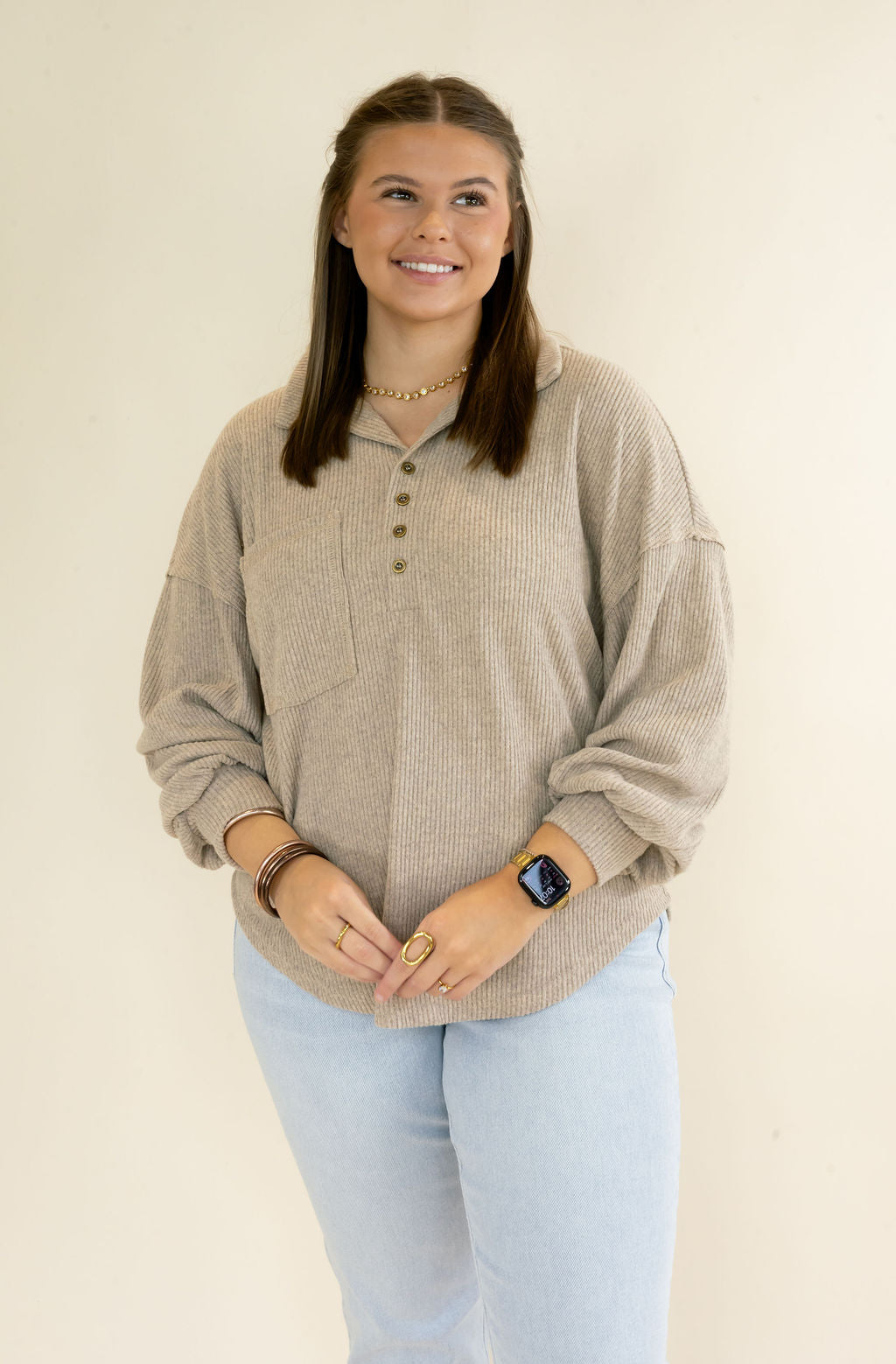 A woman modeling a beige ribbed long-sleeve henley shirt with a button placket, paired with light blue jeans, against a plain cream background.
