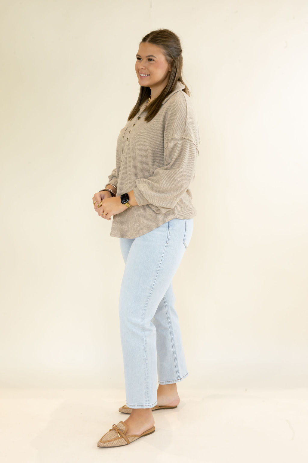 A woman modeling a light beige long-sleeve knit top and light blue jeans, standing in profile against a plain off-white studio background.