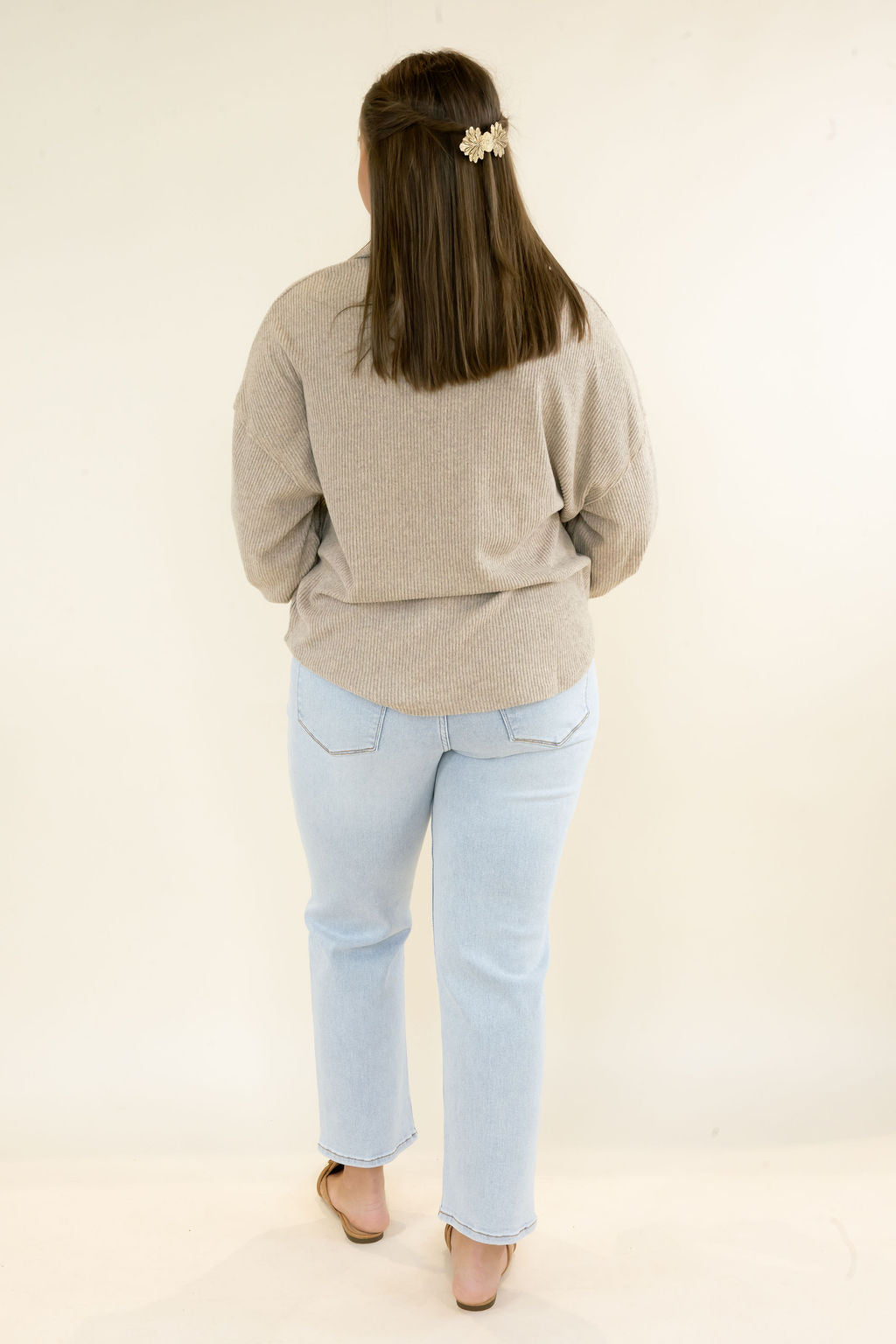 A woman viewed from behind wearing a light beige knit sweater and light blue jeans, standing against a plain off-white studio background.
