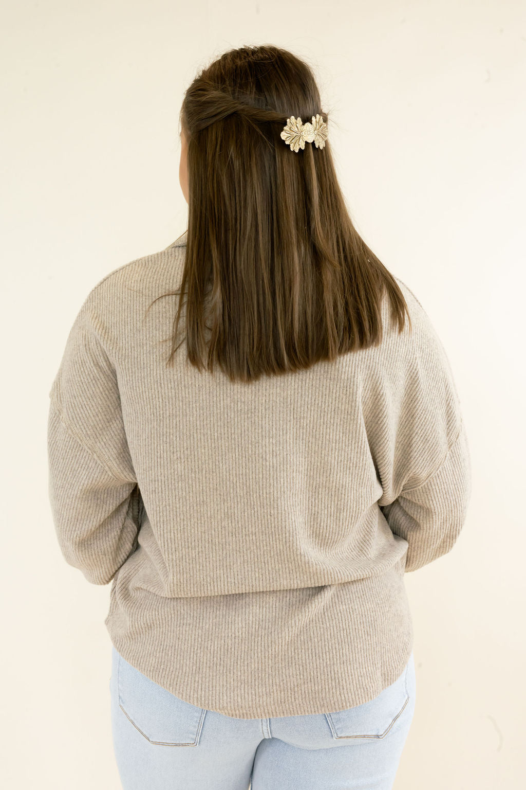 A woman viewed from behind wearing a beige ribbed knit sweater and light blue jeans, with a small cream floral hair clip holding back her brown hair against a plain off-white background.
