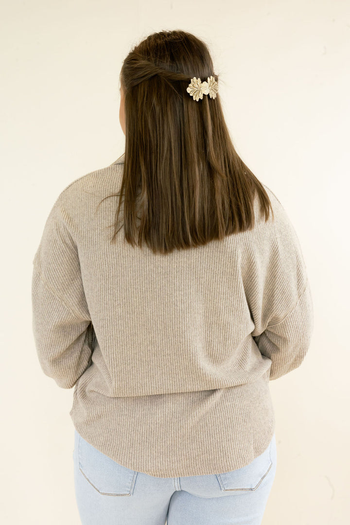 A woman viewed from behind wearing a beige ribbed knit sweater and light blue jeans, with a small cream floral hair clip holding back her brown hair against a plain off-white background.