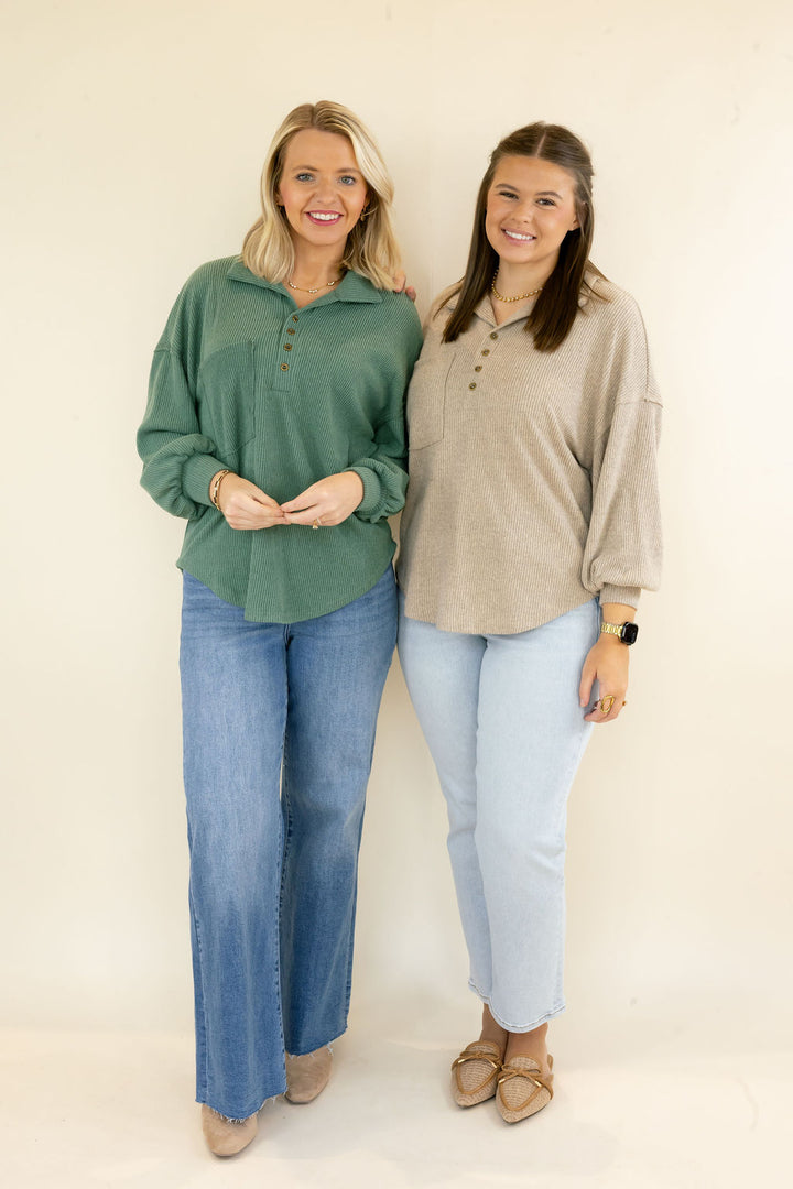 Two women standing side-by-side modeling long-sleeve button-up henley shirts. The woman on the left wears a sage green shirt with blue jeans, and the woman on the right wears a light taupe/beige shirt with light-wash blue jeans.