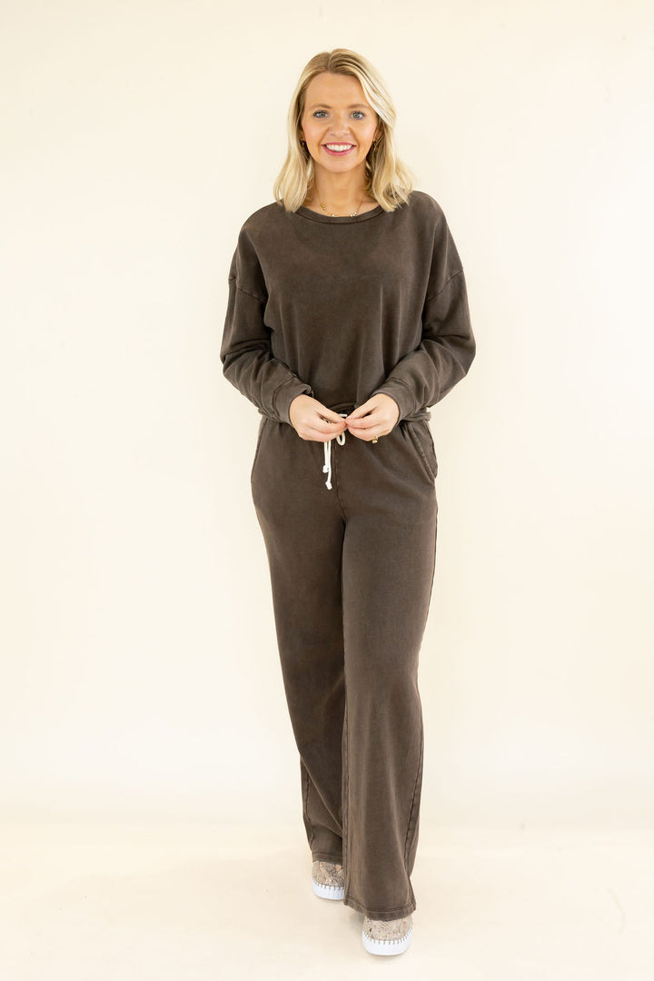 A woman modeling a matching two-piece lounge set consisting of a dark brown long-sleeve top and wide-leg pants, standing against a plain off-white studio background.