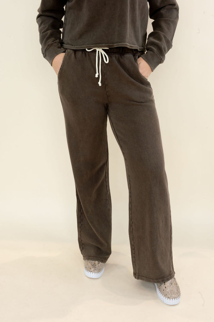 A person wearing dark brown wide-leg sweatpants with a white drawstring, paired with a matching dark sweatshirt and gray slip-on shoes, standing against a plain off-white studio background.
