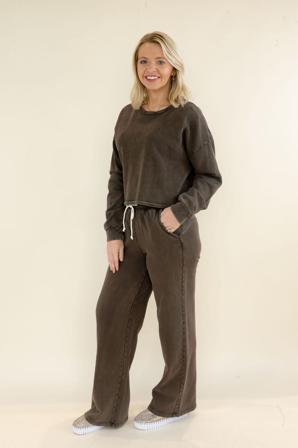 A woman modeling a dark brown lounge set consisting of a long-sleeve cropped sweatshirt and matching wide-leg drawstring pants, standing against a plain beige studio background.