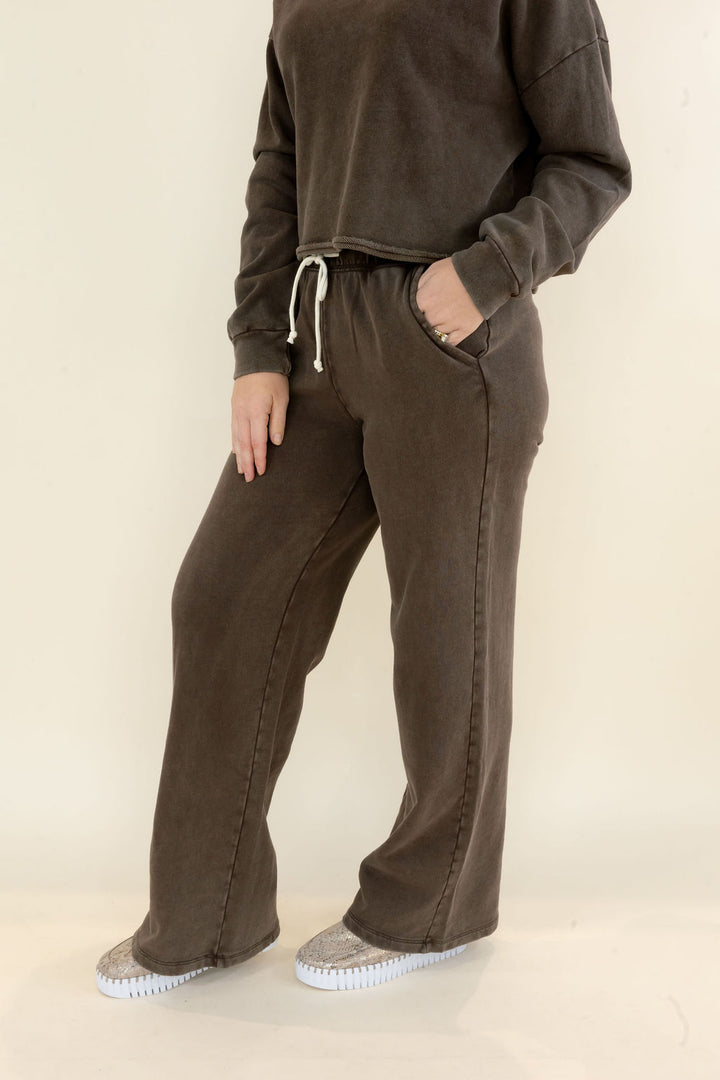 A person wearing a matching dark brown lounge set consisting of a cropped long-sleeve sweatshirt and wide-leg drawstring sweatpants, paired with gray slip-on shoes, against a plain off-white studio background.