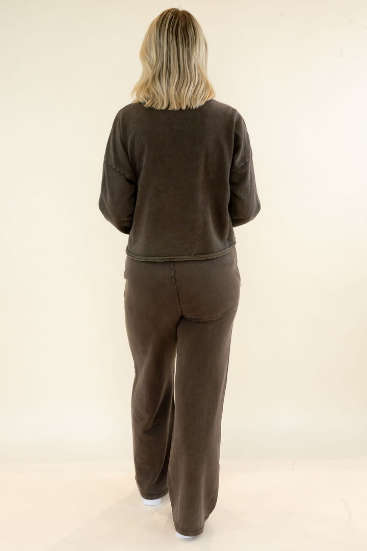 A woman viewed from behind wearing a matching dark brown lounge set consisting of a cropped long-sleeve sweatshirt and wide-leg sweatpants, set against a plain cream studio background.