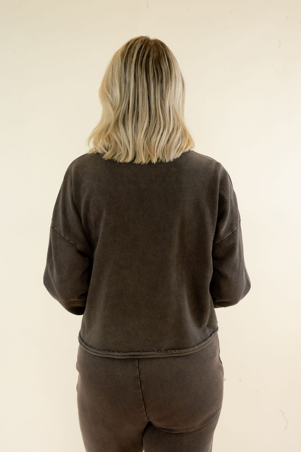 A person viewed from behind wearing a dark brown washed sweatshirt and matching sweatpants against a plain off-white background.