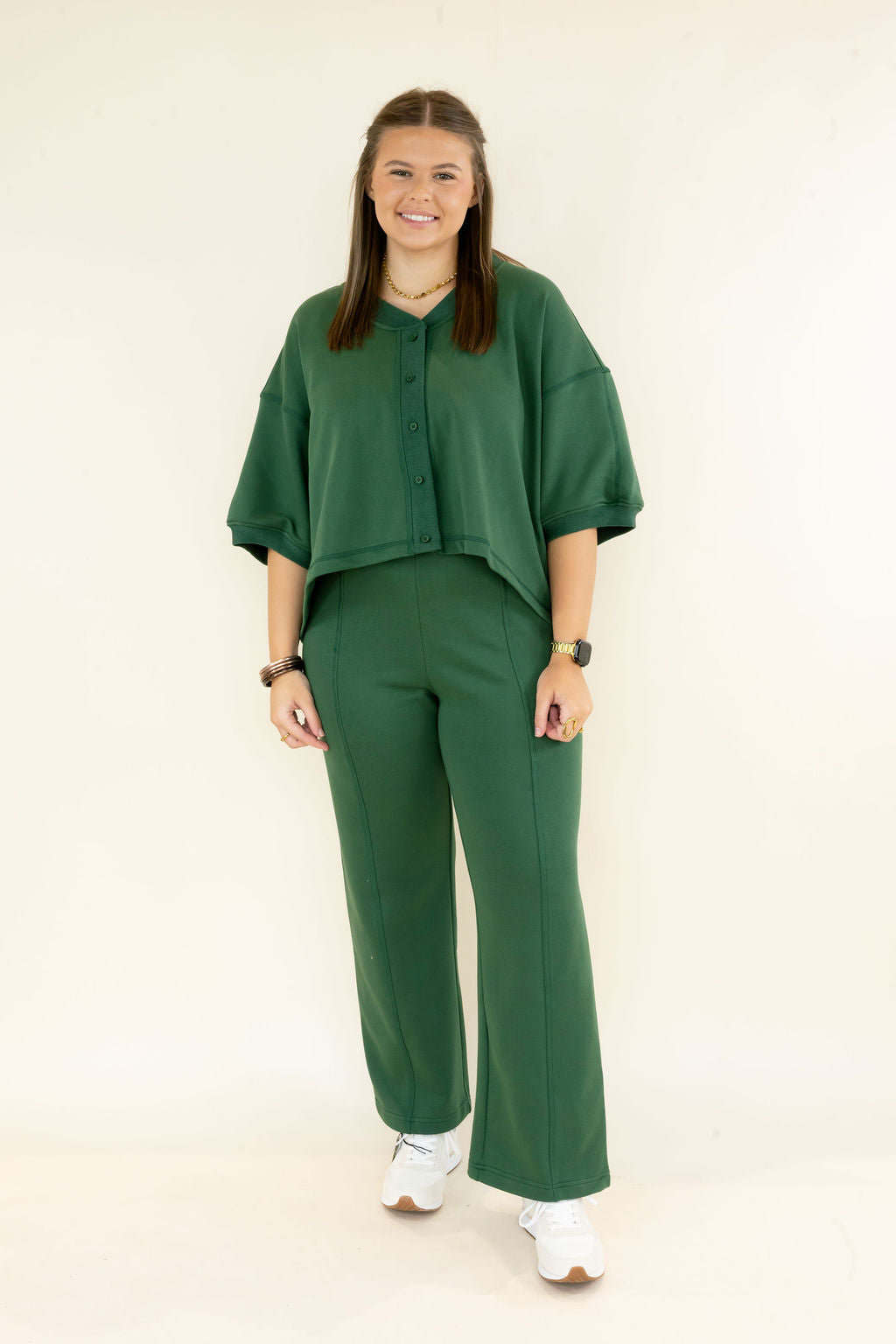 A woman modeling a matching dark green two-piece set consisting of a short-sleeve button-front top and wide-leg trousers, paired with white sneakers, against a plain off-white studio background.