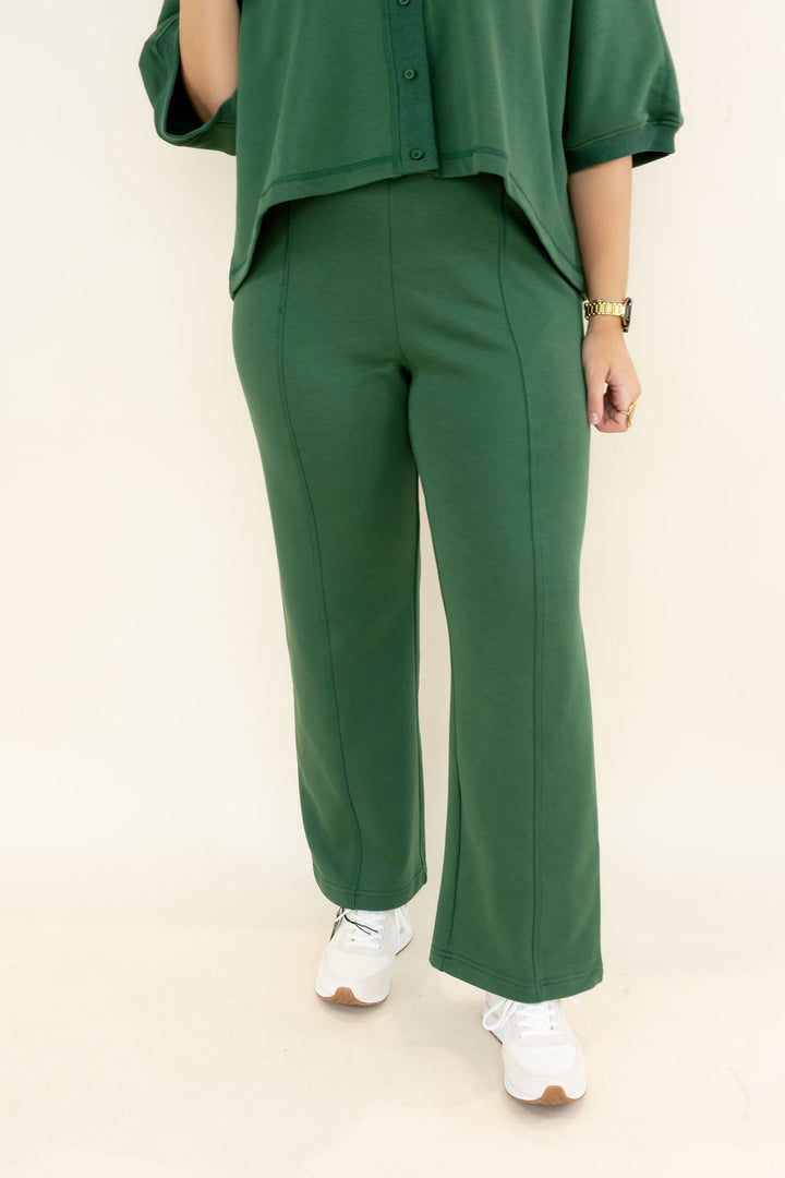 A person wearing a matching green two-piece outfit with a short-sleeve button-front top and high-waisted wide-leg pants, paired with white sneakers, photographed against a plain light background.