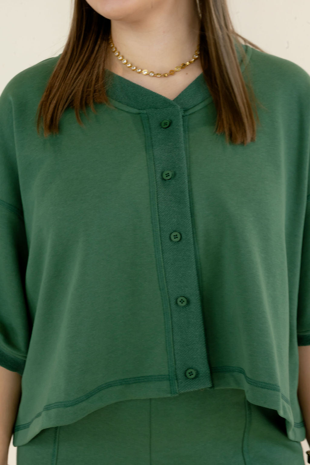 A close-up of a model wearing a dark green short-sleeve top with a buttoned front placket and a relaxed cropped fit, paired with a gold chain necklace.