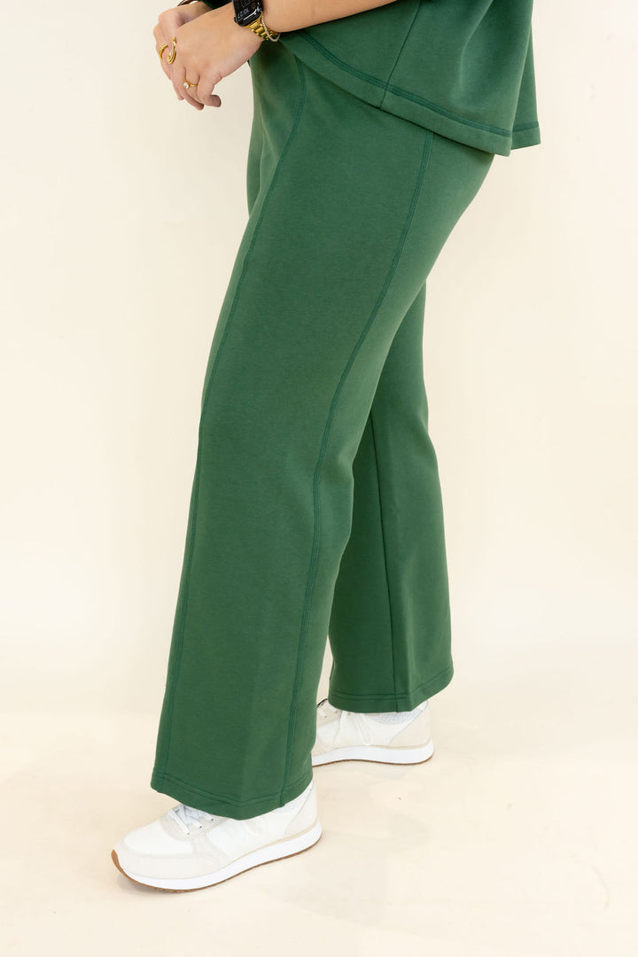 A person wearing dark green wide-leg sweatpants and white sneakers, shown from the waist down against a plain cream studio background.