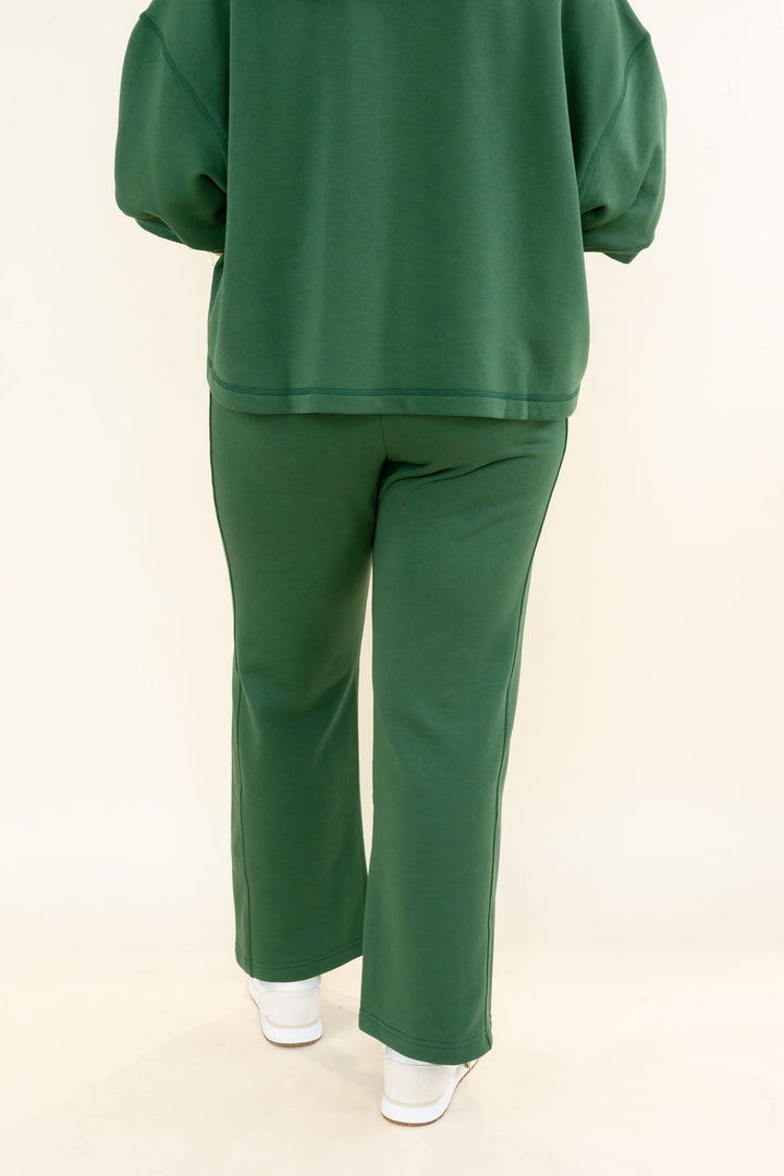 A person shown from the back wearing a matching dark green lounge set consisting of a long-sleeve sweatshirt and wide-leg pants, paired with white sneakers, against a plain off-white studio background.