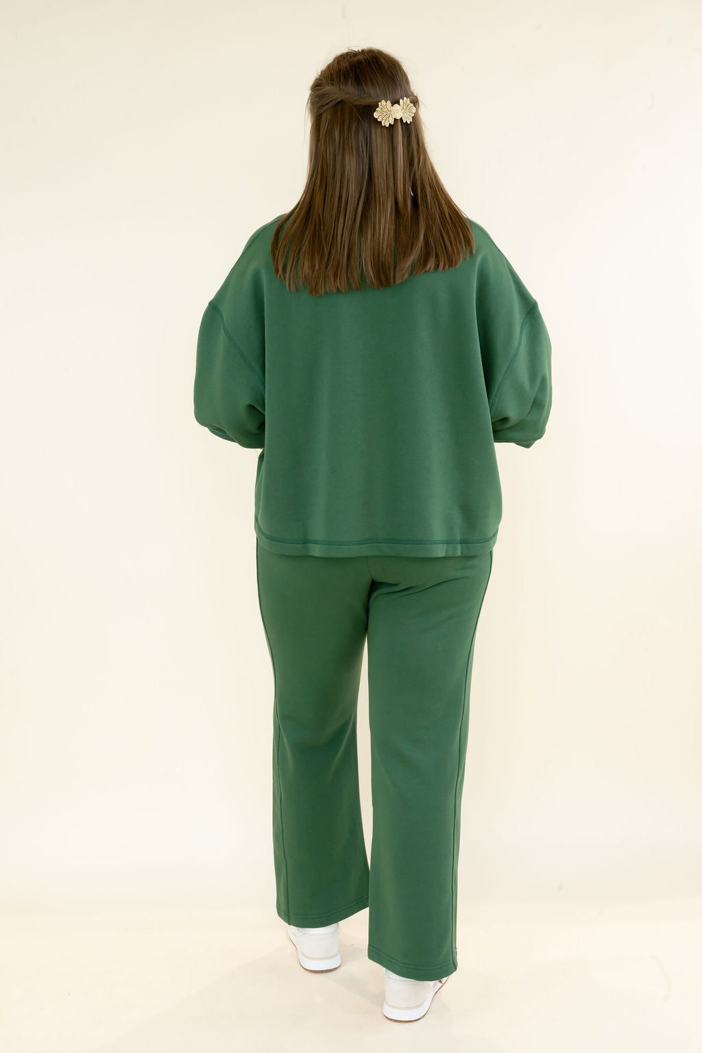 A woman viewed from behind wearing a matching green two-piece lounge set consisting of a cropped sweatshirt with balloon sleeves and wide-leg pants, styled with white sneakers against a plain cream background.