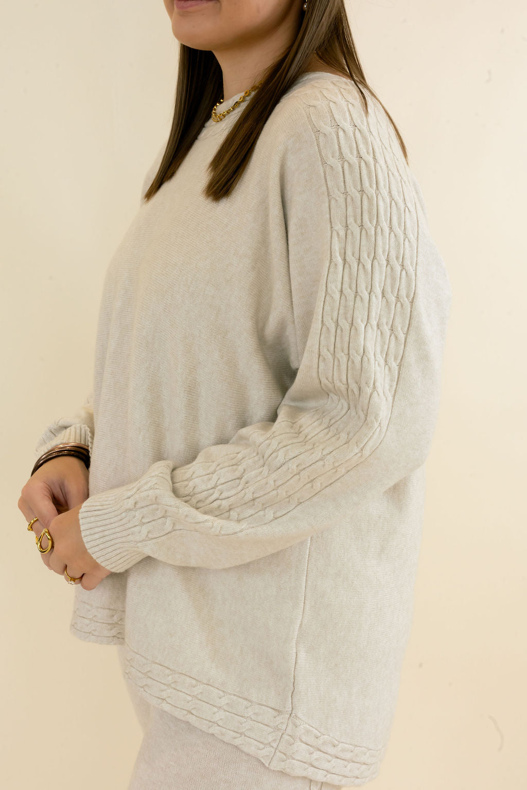 Angled close-up of light oatmeal knit pullover showing textured sleeves, relaxed silhouette, side slit hem, and long sleeves