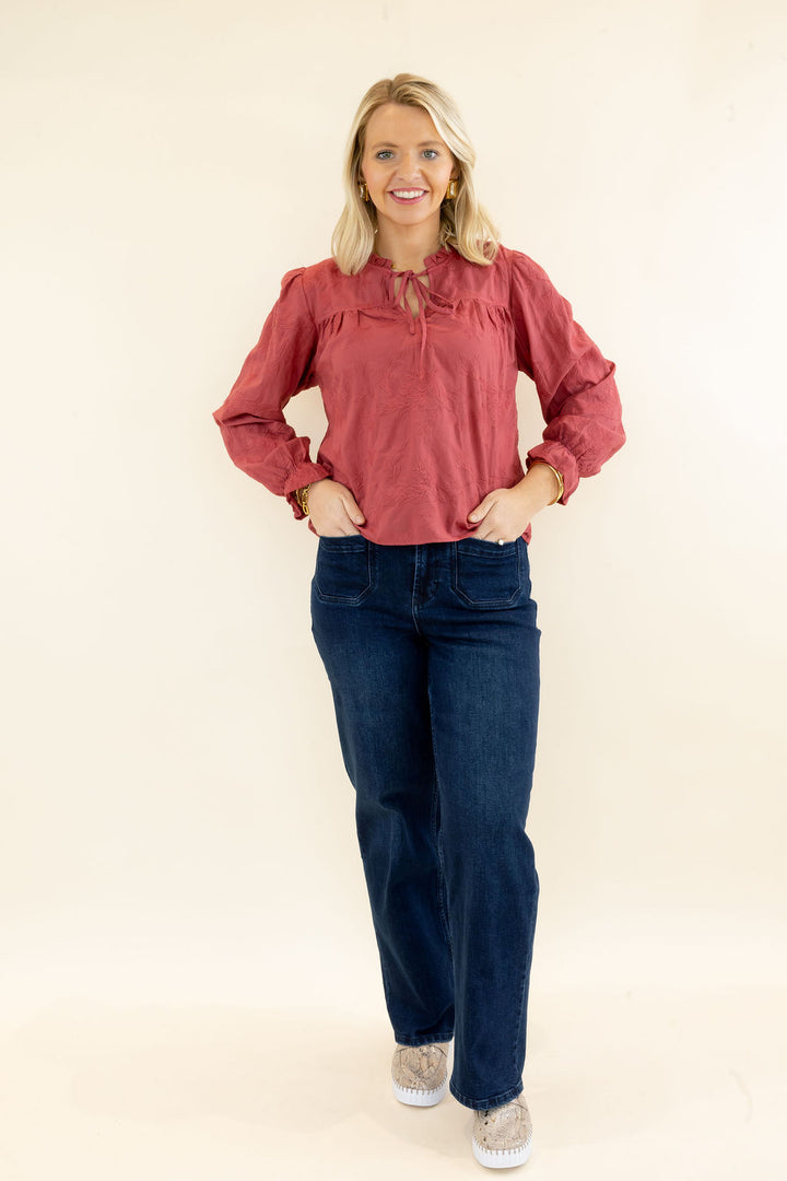 Woman modeling rust red long sleeve blouse with ruffle neckline, tie-front detail, and dark wash wide leg jeans