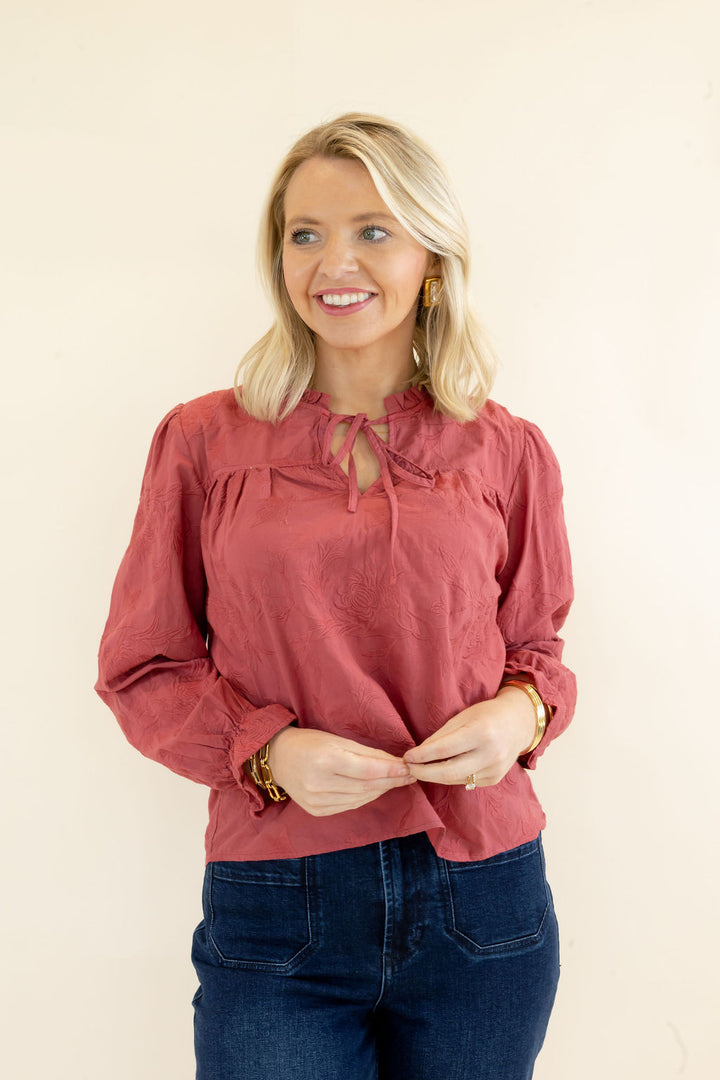 Woman wearing rust red ruffle neck blouse with tie detail, long sleeves, and relaxed fit styled with dark wash jeans