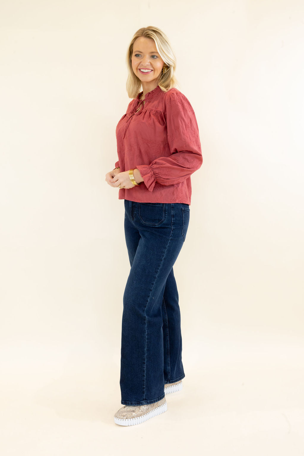 Side view of woman modeling rust red ruffle neck blouse with relaxed fit and dark wash wide leg jeans