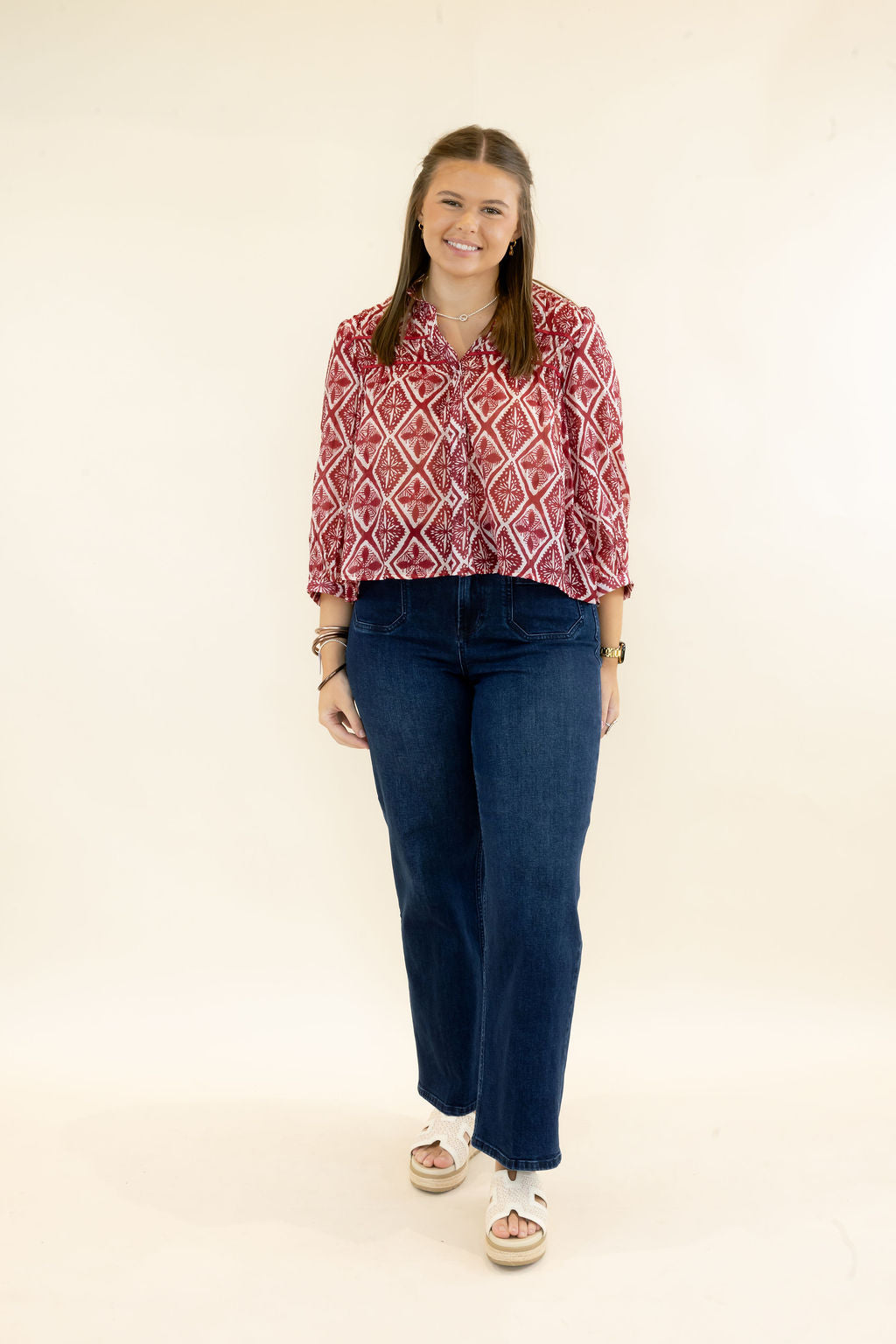 Full-length view of woman wearing red and ivory printed blouse with relaxed fit styled with dark wash wide leg jeans and neutral sandals