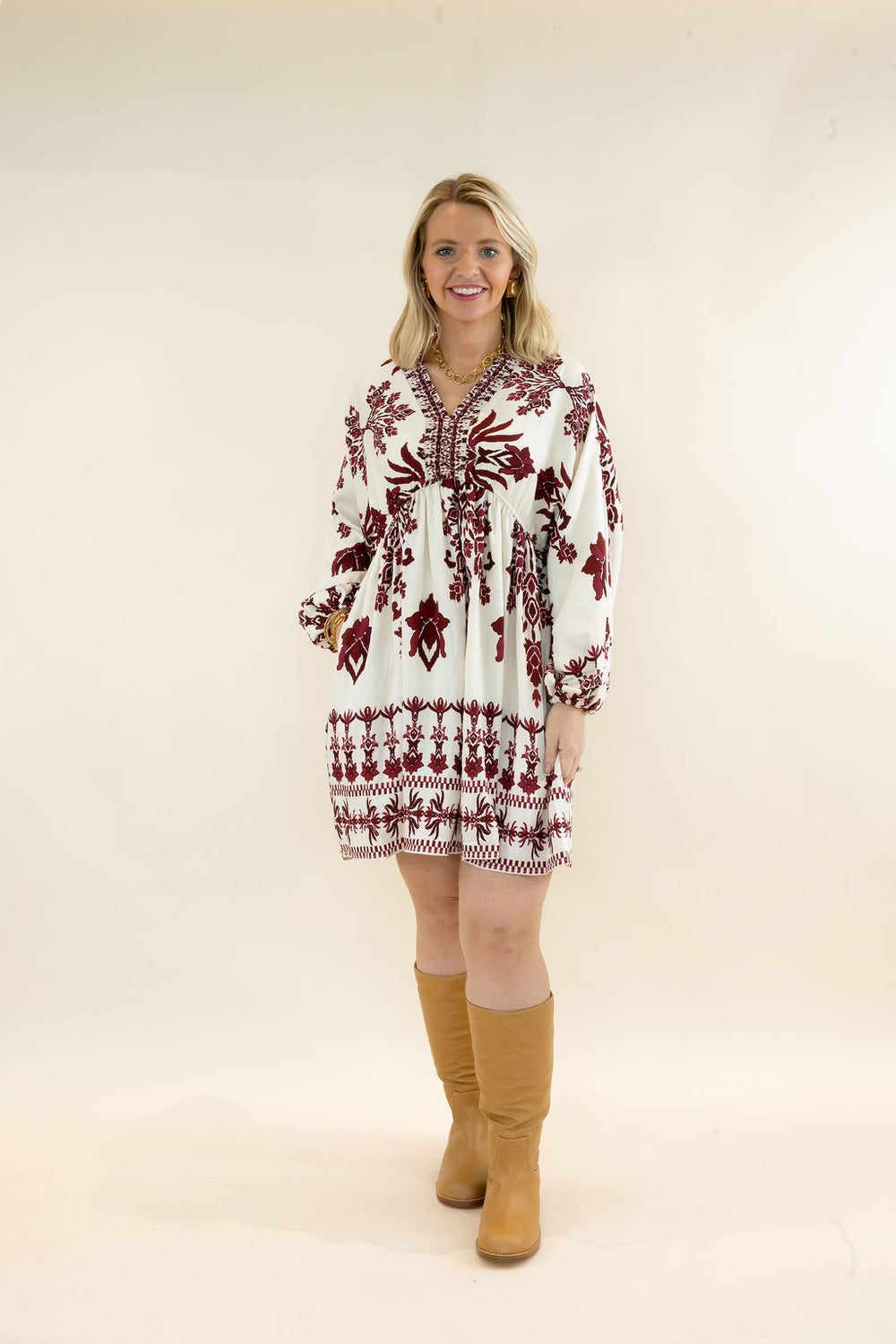 Full-length view of woman wearing ivory and burgundy floral print mini dress with long sleeves and tan knee-high boots