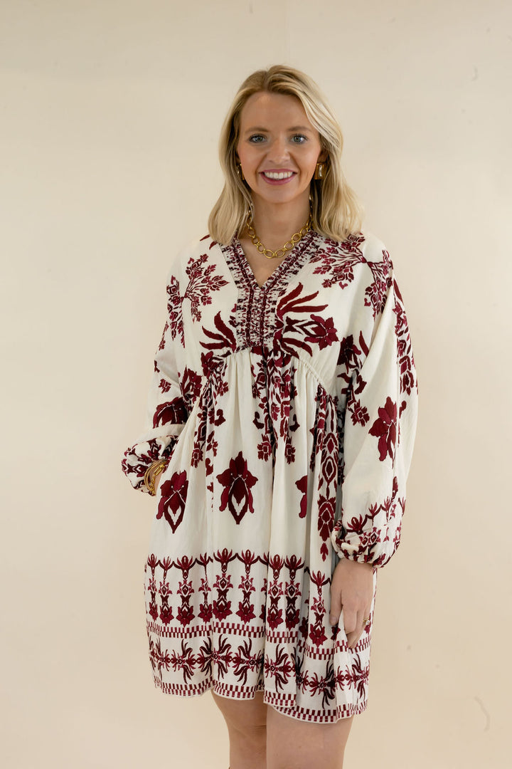 Woman modeling ivory mini dress with burgundy botanical print, V-neckline, long balloon sleeves, gathered waist, and decorative border hem
