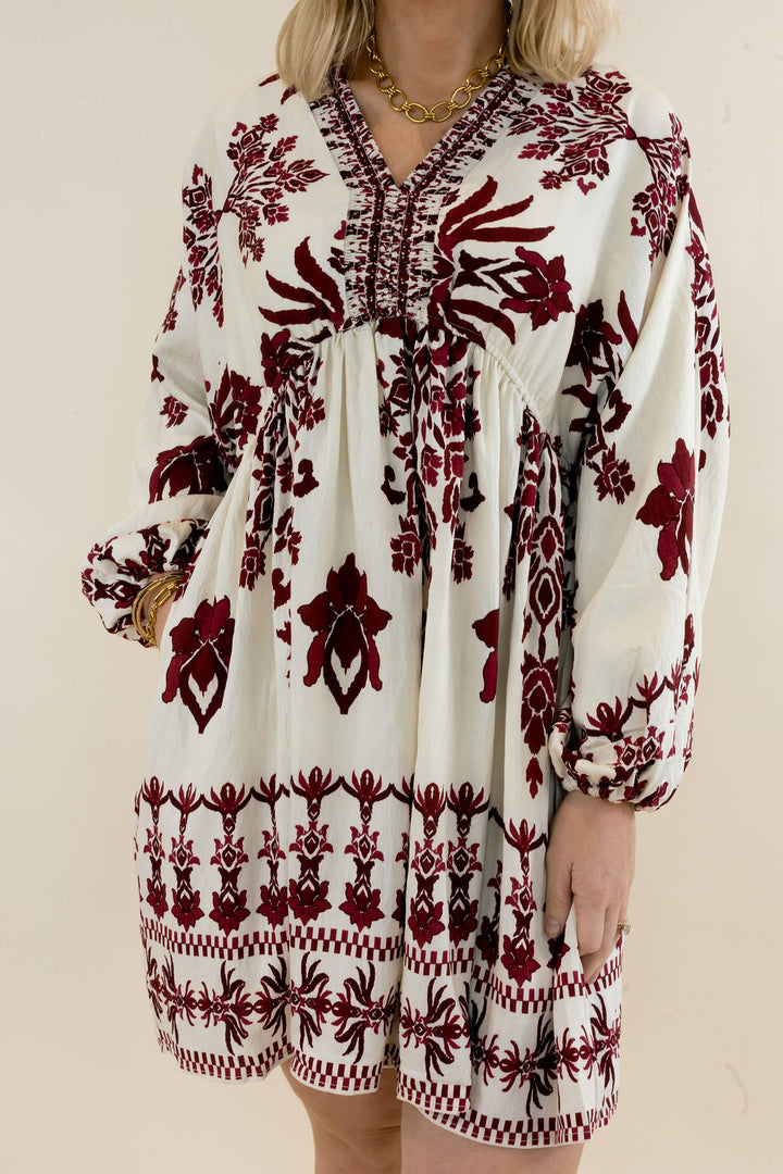 Close-up of ivory mini dress highlighting the burgundy botanical print, V-neckline, decorative trim, gathered waist, and long balloon sleeves