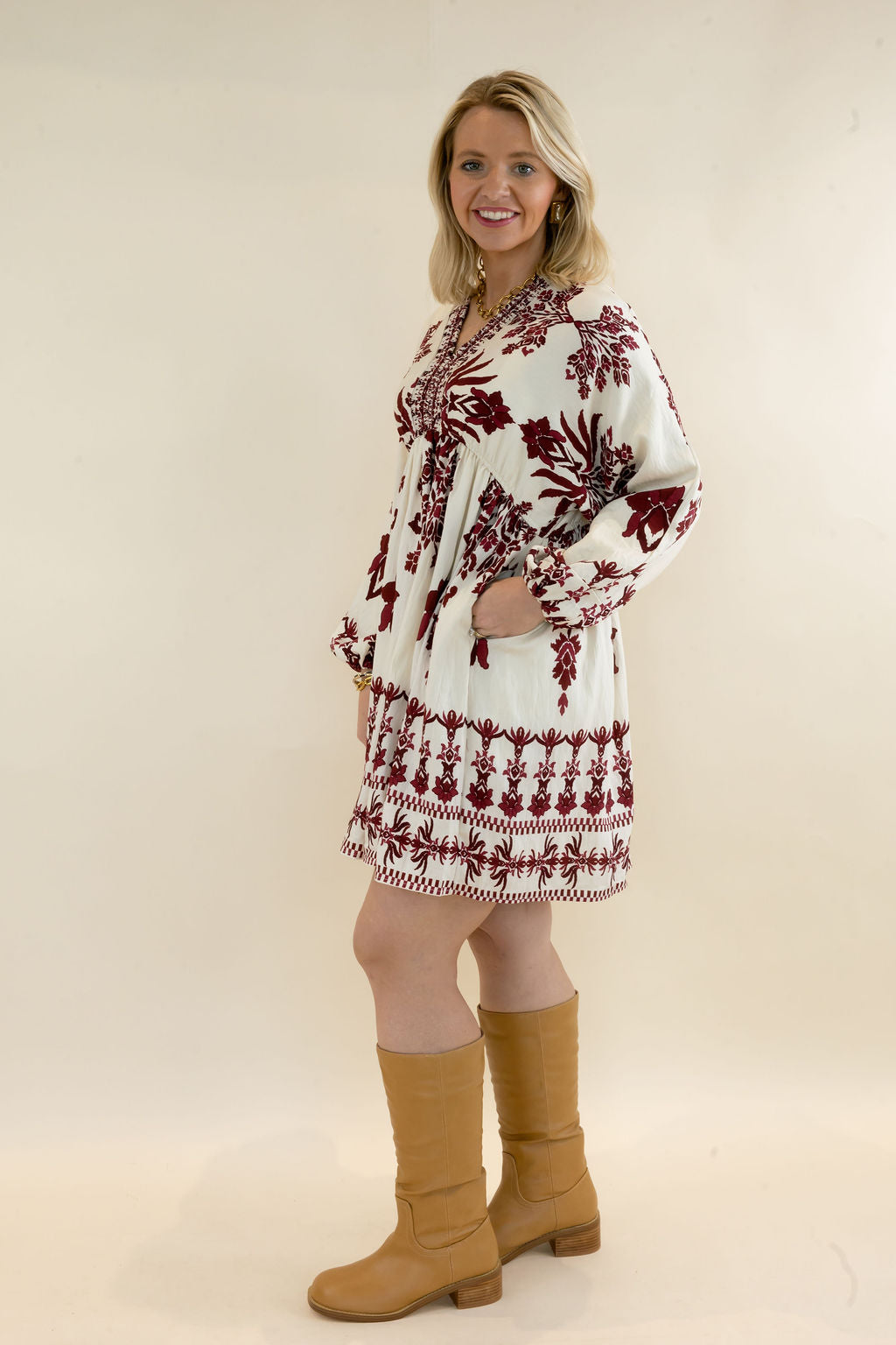 Side view of woman modeling ivory and burgundy printed mini dress with flowy silhouette, long sleeves, and tan knee-high boots