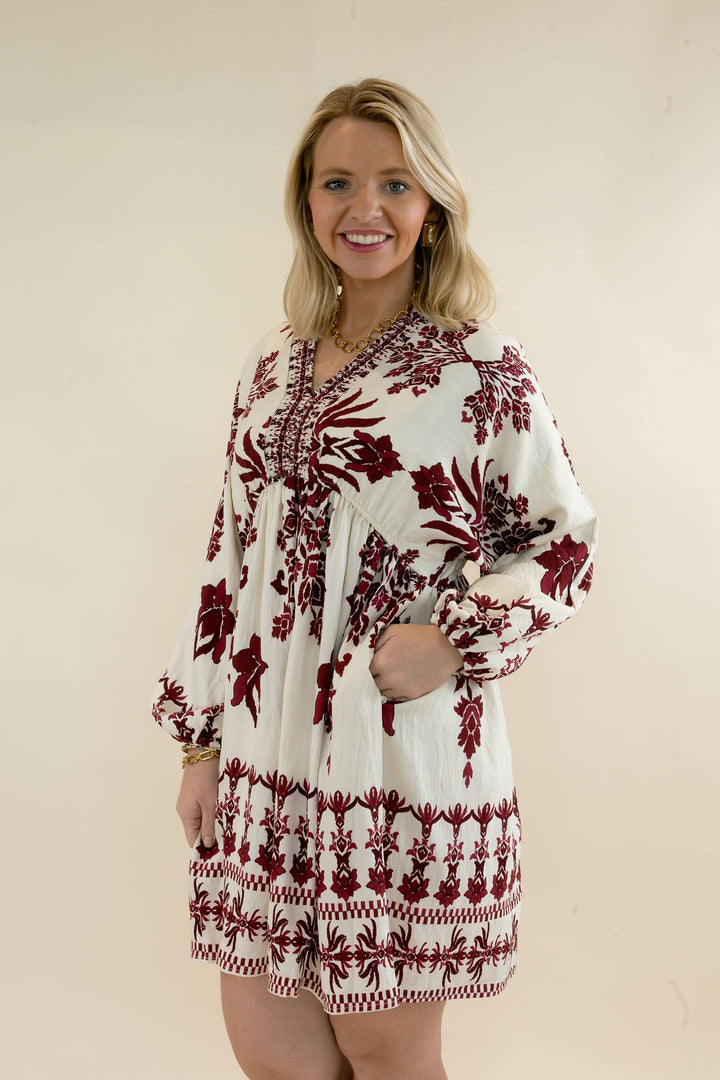 Three-quarter view of ivory and burgundy botanical print mini dress featuring V-neckline, gathered waist, long balloon sleeves, and border print hem