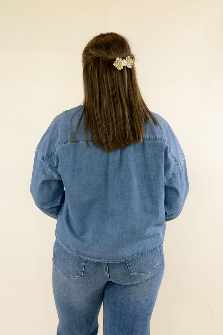 Close back view of light wash denim button-up shirt highlighting the relaxed silhouette, long sleeves, back seam detail, and curved hem
