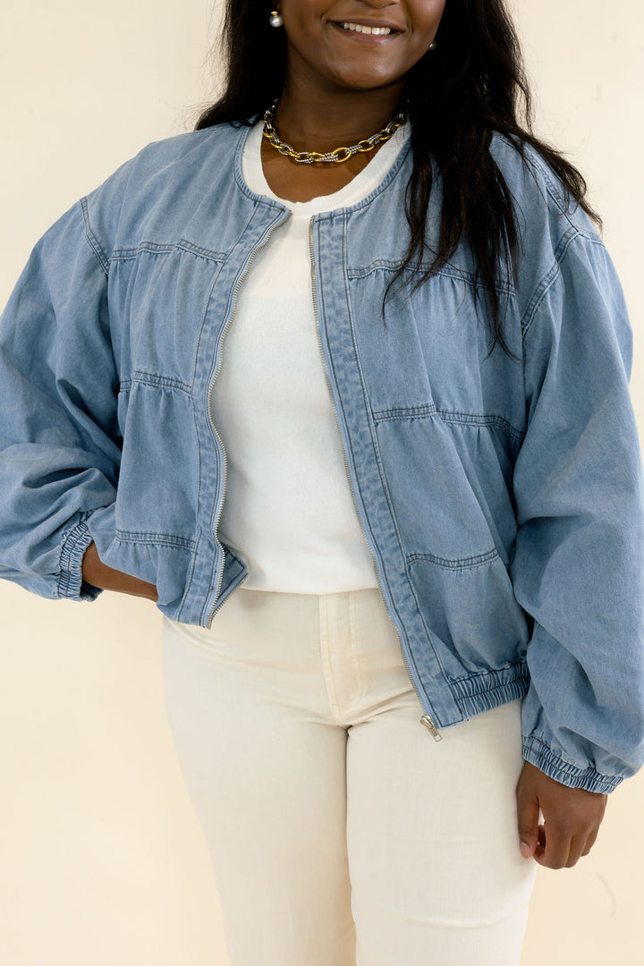Close-up of light wash denim bomber jacket showing snap button closure, gathered seam detailing, balloon sleeves, and elastic hem