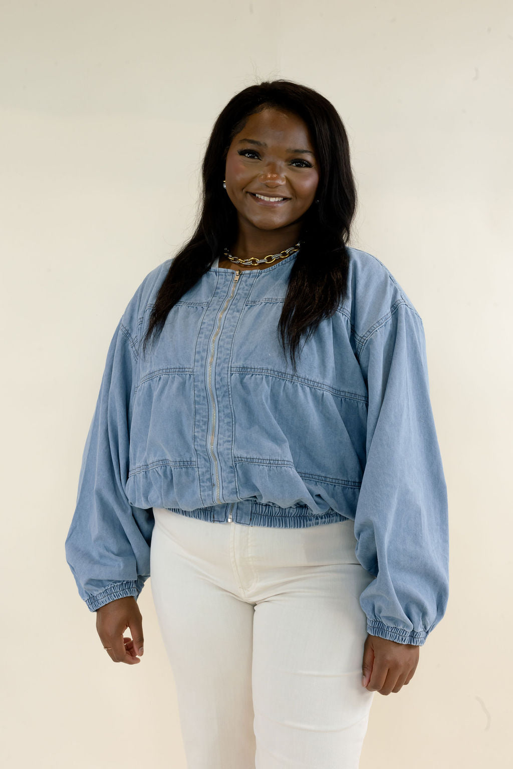 Woman modeling light wash denim bomber jacket with round neckline, balloon sleeves, gathered elastic hem, and white pants