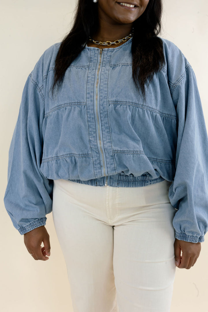 Front view of light blue denim bomber jacket highlighting the snap button closure, gathered bodice, long balloon sleeves, and elasticized hem