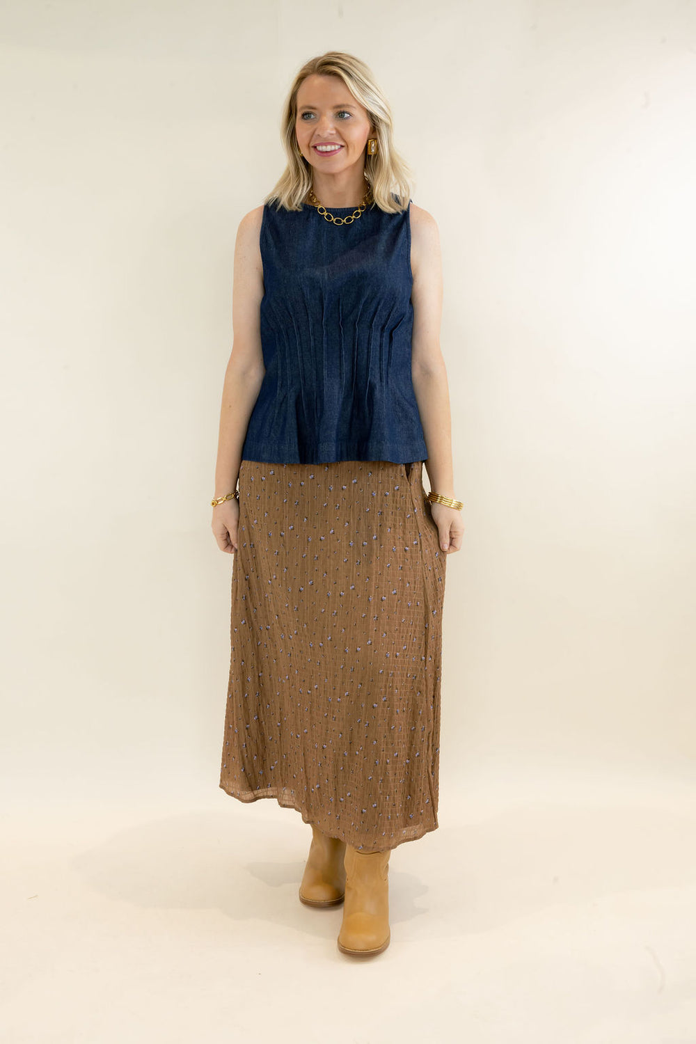 Full-length view of woman wearing dark indigo sleeveless denim peplum top styled with a brown printed midi skirt and tan boots