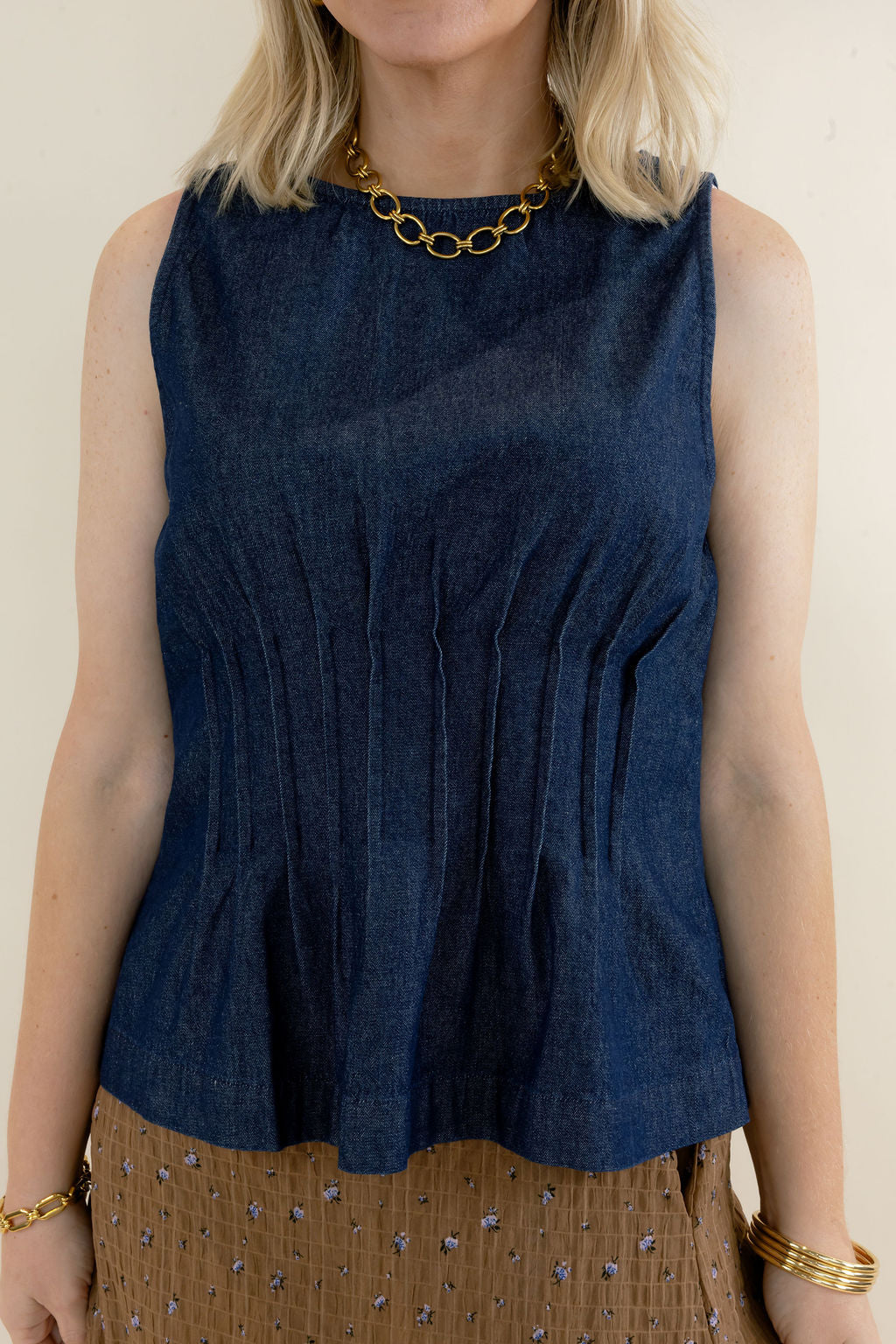 Close-up of dark indigo sleeveless denim top highlighting the round neckline, vertical pleat detailing, fitted waist, and flared peplum hem