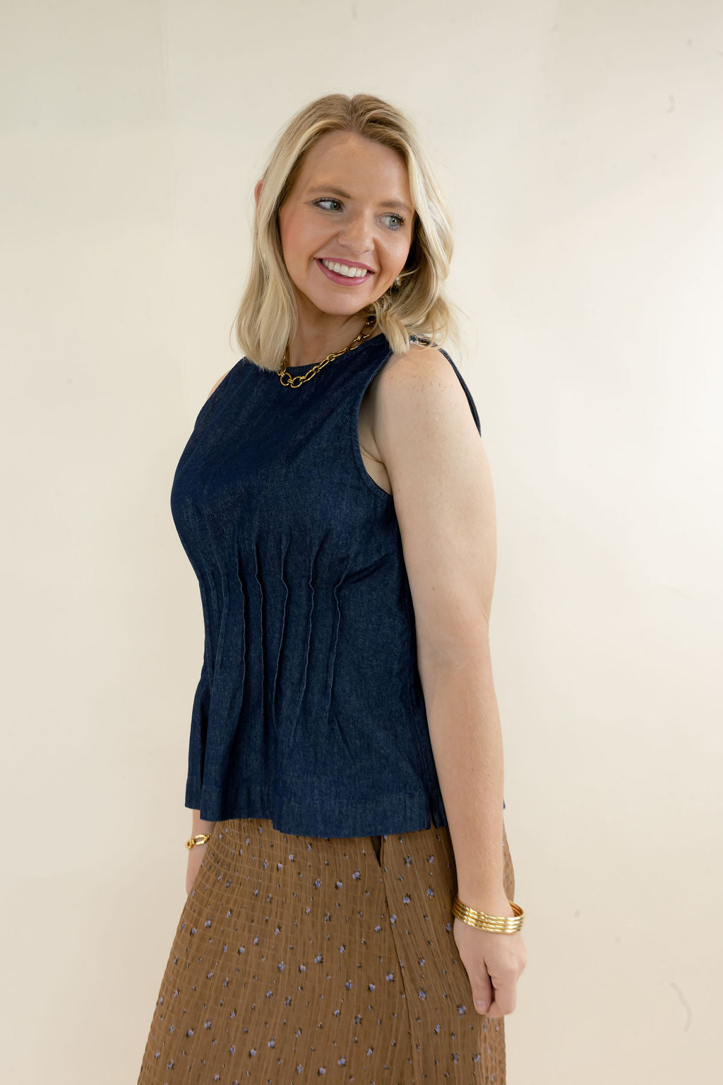 Three-quarter view of dark indigo sleeveless denim top featuring a fitted pleated bodice, round neckline, and flared peplum silhouette