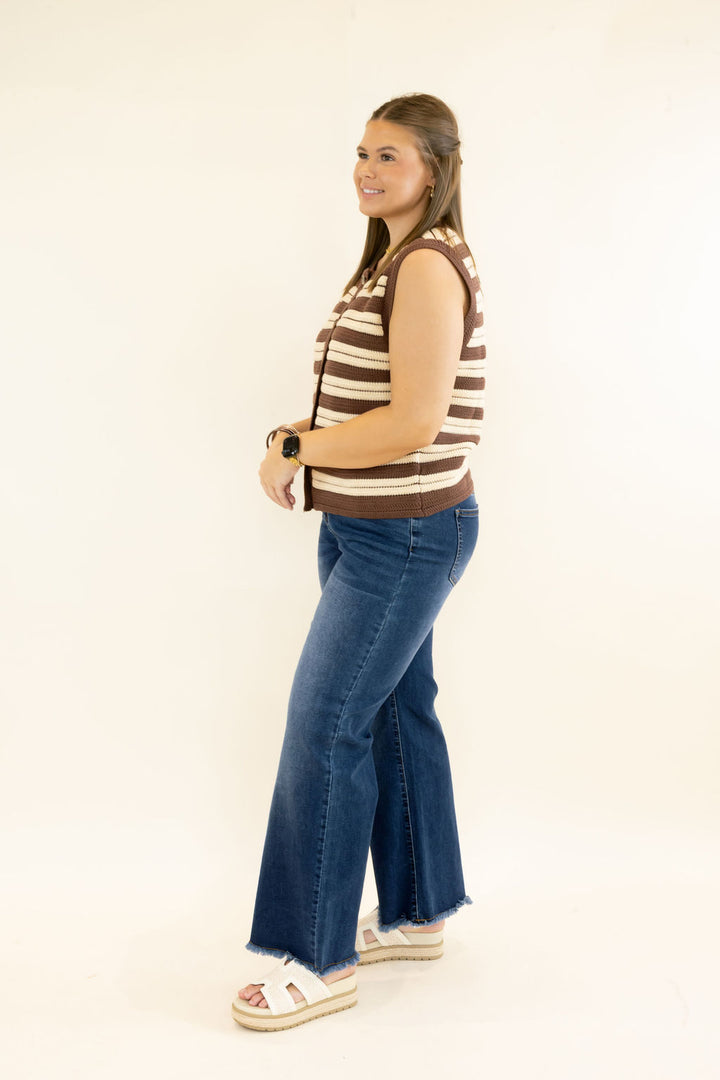 Side view of woman modeling brown and cream striped sleeveless sweater vest with button front and dark wash wide leg jeans