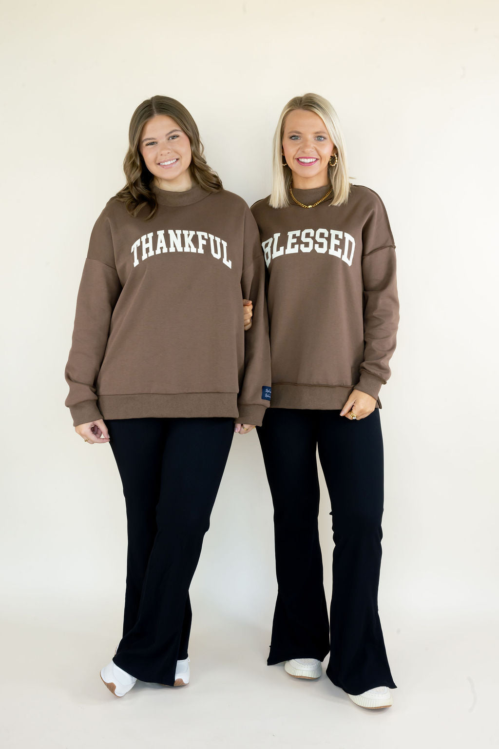 Two women modeling matching brown crewneck sweatshirts with white arched text, one reading &