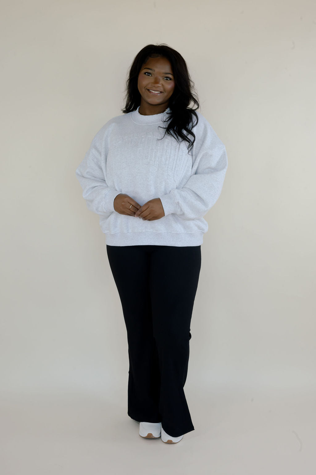 Full-length view of woman wearing heather gray Simply For Good crewneck sweatshirt with black flare pants and white shoes